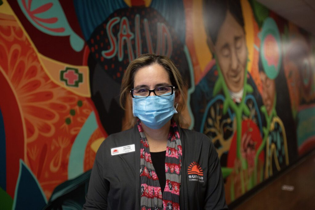 Photo of a Vitamin Angels program partner employee standing in front of a decorative, colorful wall. The person wears black glasses and a blue mask.