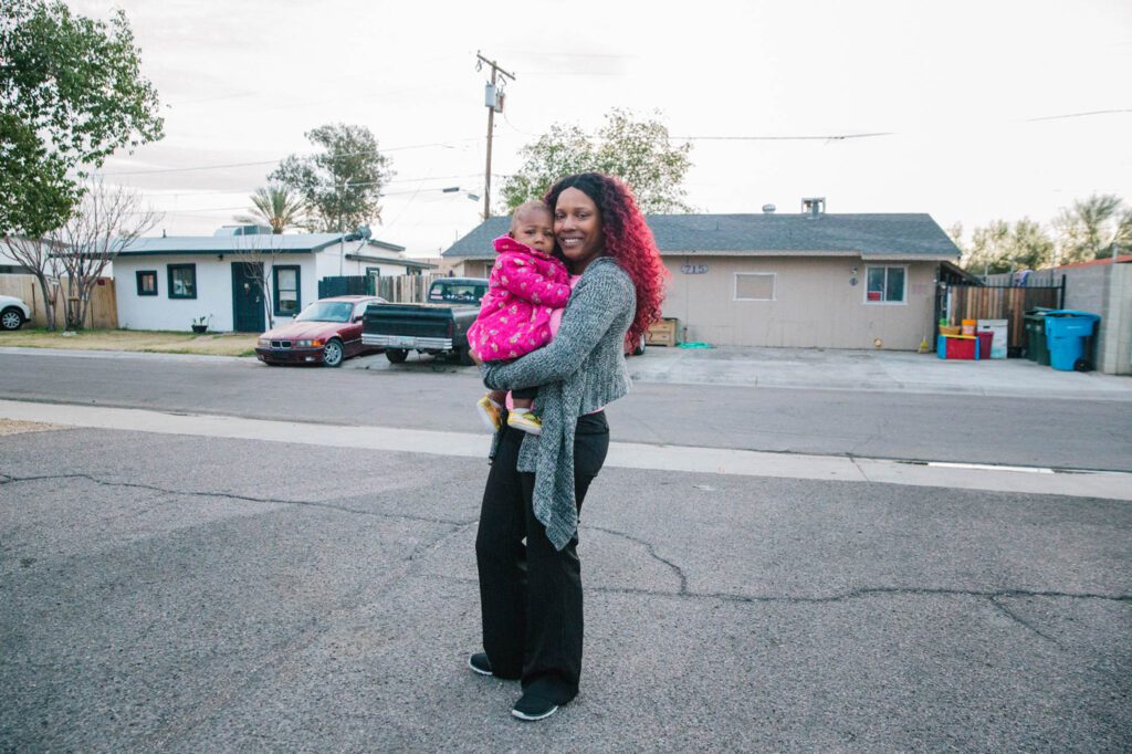 A woman of color with pink dyed hair smiles while holding young child in pink jacket in Arizona.